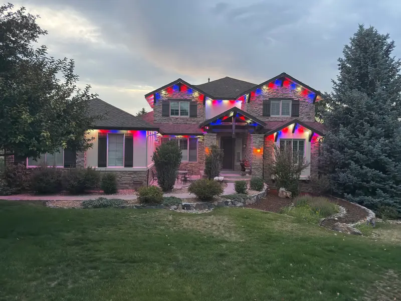 Whole-house view from the street of a stone-and-stucco Denver Metro home at dusk with Govee Pro permanent lights in red, white, and blue on every gable